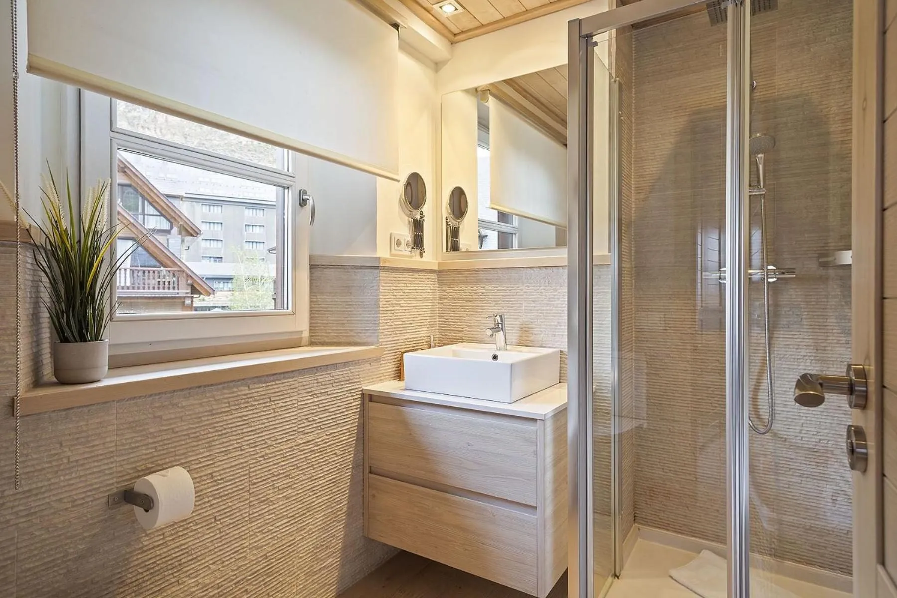 Un baño con lavabo sobre mueble de madera, ventana y ducha de cristal