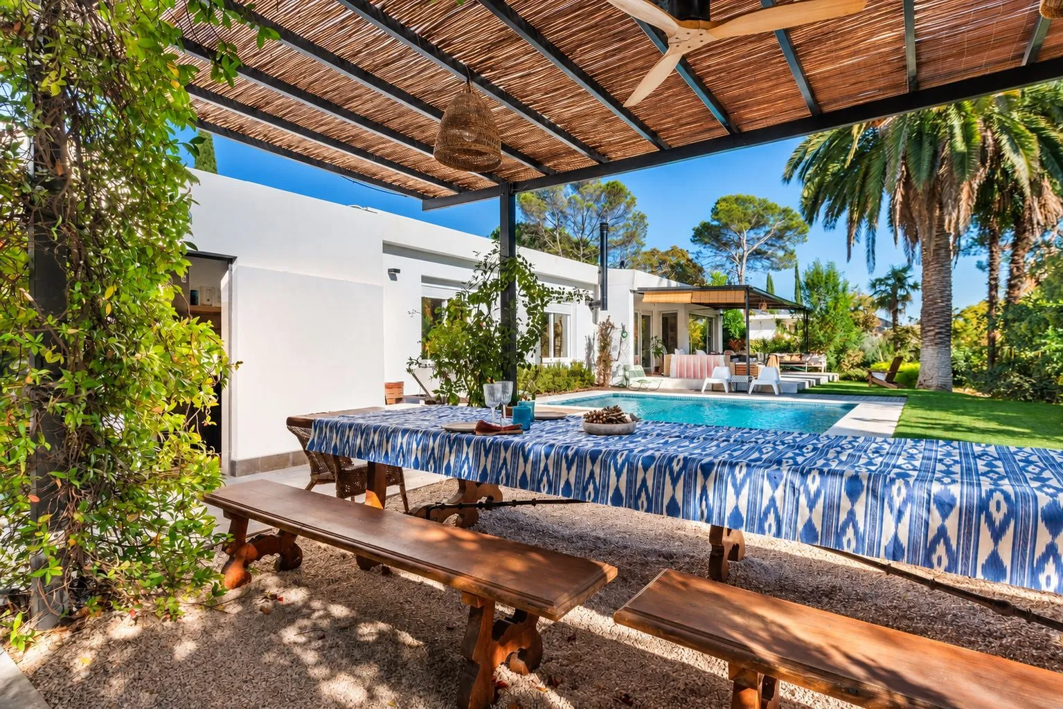 Una mesa larga con bancos de madera bajo la pérgola, con la piscina detrás