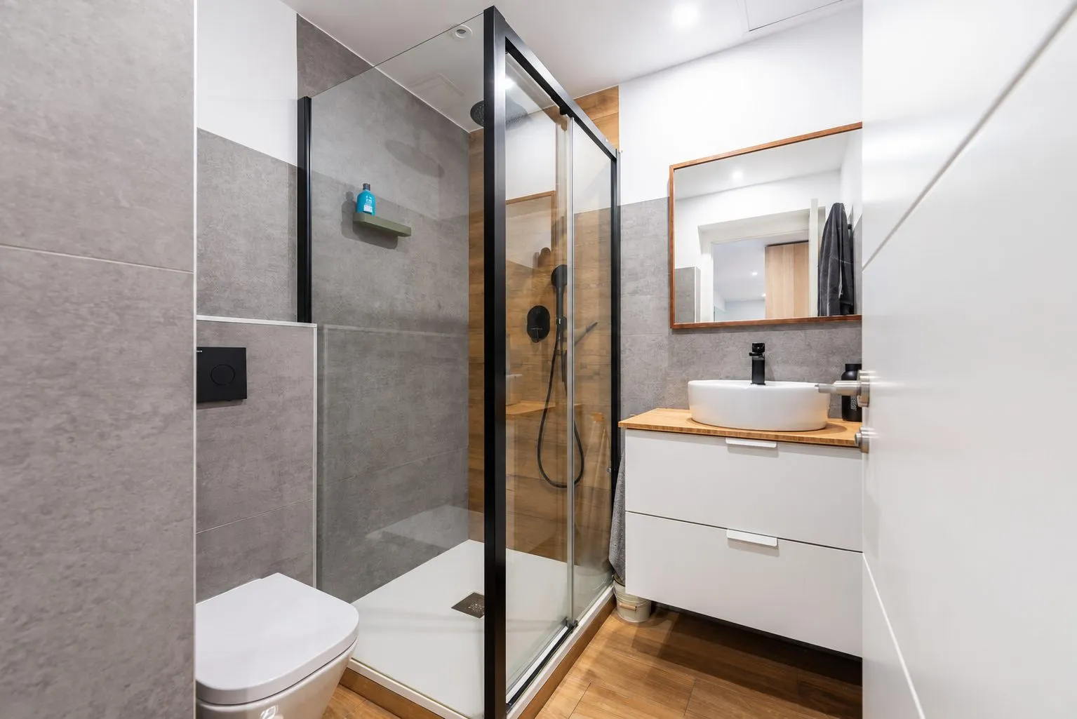 Un baño con mampara negra, lavabo sobre mueble blanco y espejo de madera