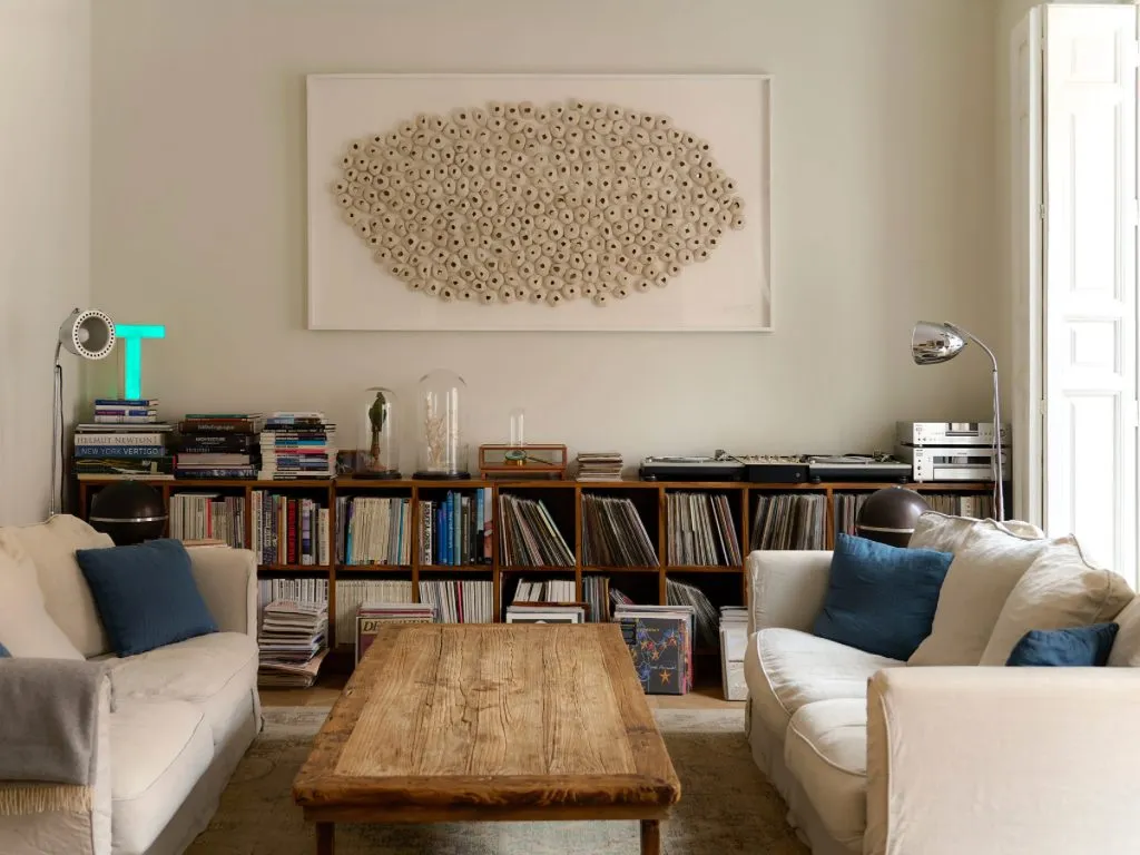 Un salón con dos sofás blancos enfrentados, cojines azules, una mesa baja de madera vieja y una estantería baja llena de libros y discos bajo un gran cuadro blanco