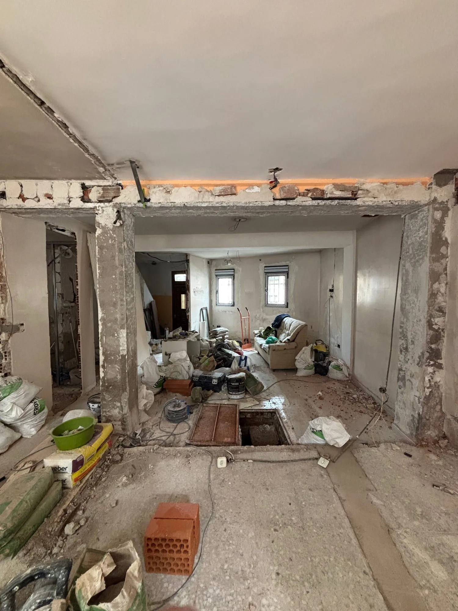 Una casa en plena obra: tabiques derribados, una viga con el ladrillo a la vista, sacos, herramientas y una trampilla abierta en el suelo